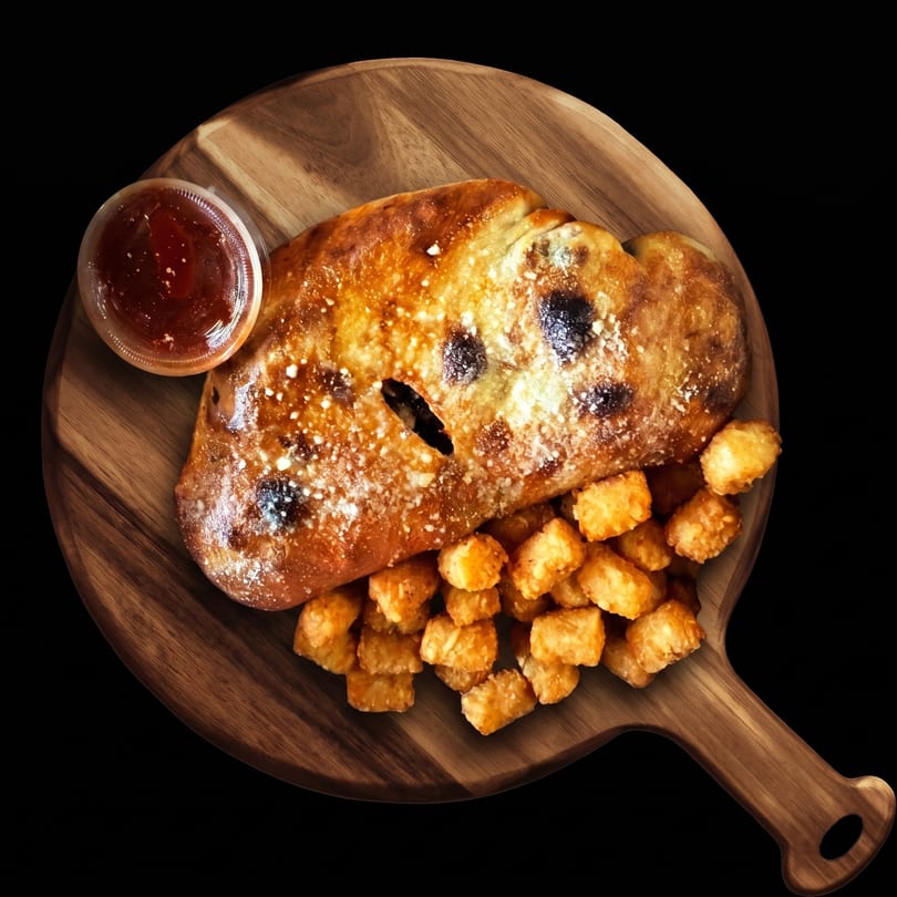 Golden-brown calzone with tater tots and a side of dipping sauce on a wooden serving board.