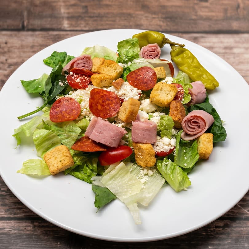 A vibrant salad with lettuce, pepperoni, ham, croutons, feta cheese, tomatoes, and a pepperoncini.